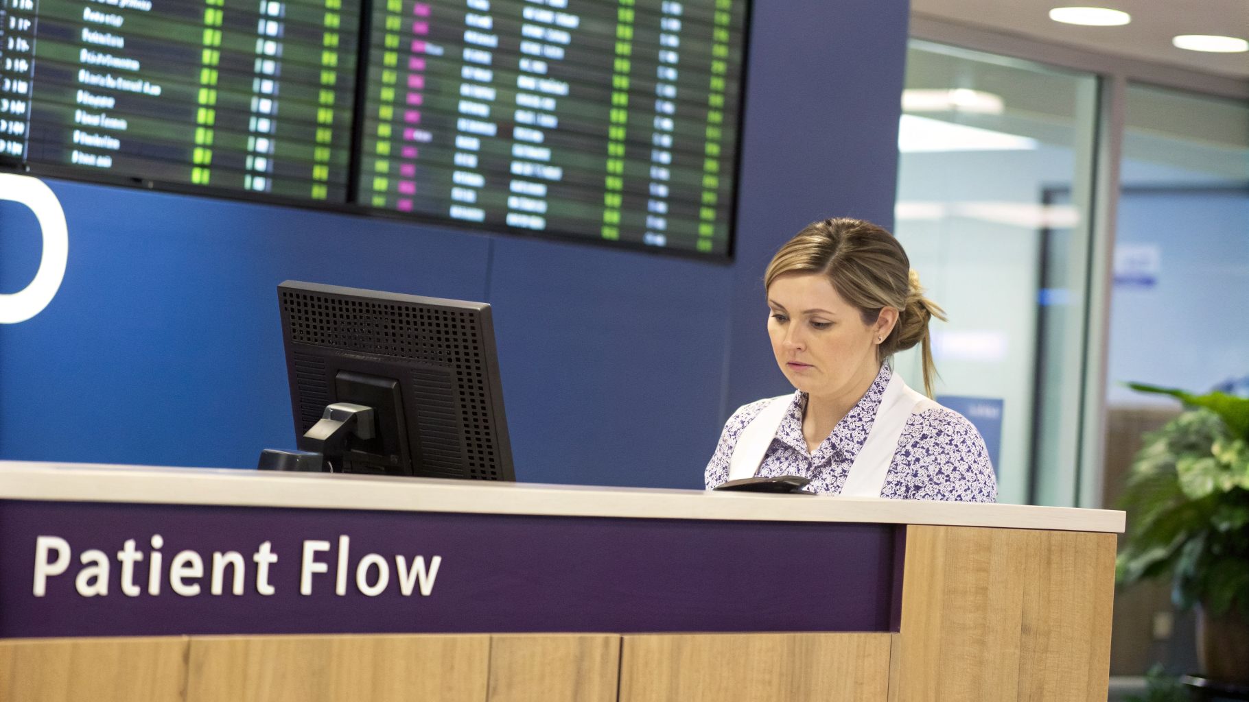 A focused female medical receptionist works at a 'Patient Flow' desk with monitors displaying patient information.